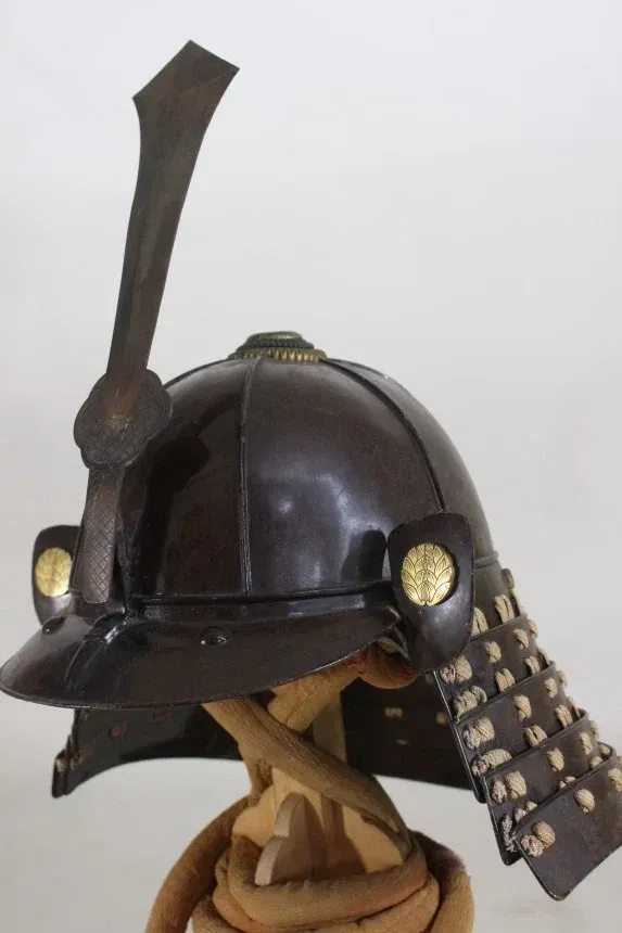 The helmet in profile, showing the brown-lacquered bowl, the tall ken maedate and a gilt crest on the turnback