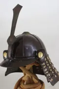The helmet in profile, showing the brown-lacquered bowl, the tall ken maedate and a gilt crest on the turnback