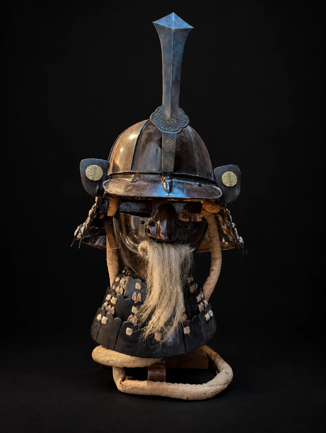 The helmet and face armour displayed together on their stand: the tall ken maedate, the gilt crests on the turnbacks, the grey moustache and the laced throat guard, with the cream cord looped at the base